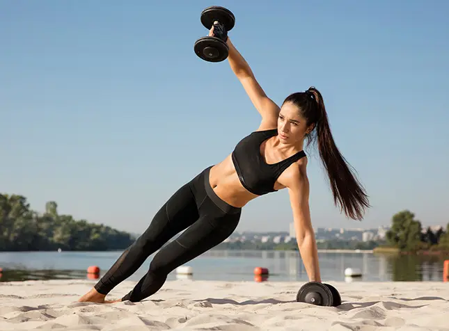 Mulher treinando com halter na areia, em um dia de sol